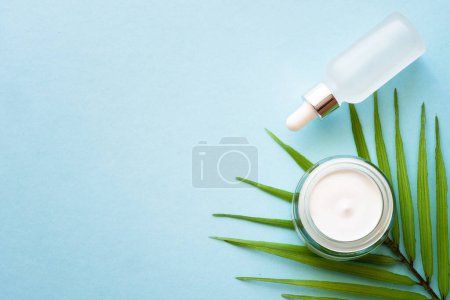 Photo for Cream jar and serum bottle with palm leaf on blue background. Natural cosmetic concept. Flat lay with copy space. - Royalty Free Image