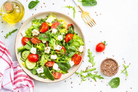 Téléchargez les photos : Salade de printemps dans une assiette artisanale. Épinards, roquette, tomates et feta à l'huile d'olive. Vue de dessus sur blanc. - en image libre de droit