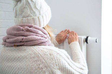 Foto de Una mujer se calienta las manos cerca de la batería. Mujer vestida cálidamente fría en la casa. Ahorro de calefacción. Facturas de servicios públicos caros. - Imagen libre de derechos