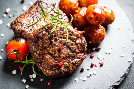 Photo for BBQ lunch. Meat steaks with potato and tomatoes at black board. Close up. - Royalty Free Image