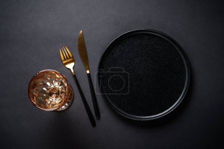 Téléchargez les photos : Table de fête avec assiette noire, verre à vin et couverts dorés. Image de pose plate avec espace de copie au noir. - en image libre de droit