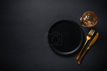 Foto de Mesa con plato negro, copa de vino y cubiertos dorados. Imagen plana con espacio de copia en negro. - Imagen libre de derechos
