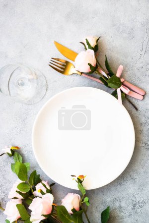 Téléchargez les photos : Assiette blanche, couverts et verre à vin aux fleurs roses. Table à ressorts. Pose plate avec espace de copie. - en image libre de droit