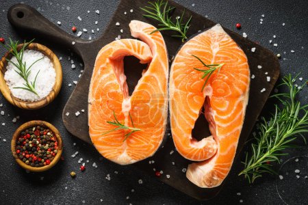 Téléchargez les photos : Steaks de saumon cru sur planche à découper à fond noir. Vue de dessus avec ingrédients pour la cuisson. - en image libre de droit