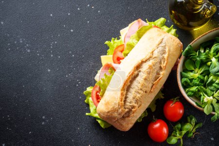 Photo for Ciabatta sandwich with lettuce, cheese, tomatoes and ham on black kitchen table with ingredients for cooking. Top view with space for text. - Royalty Free Image