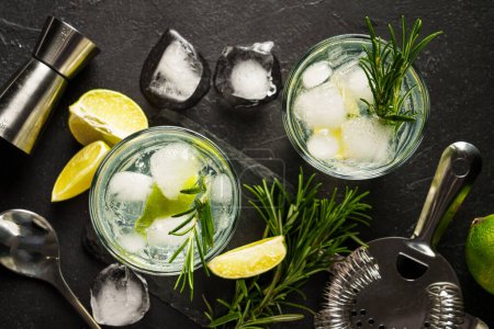 Téléchargez les photos : Gin tonique, cocktail traditionnel avec gin, tonique, glaçons, citron vert et romarin sur fond noir. Vue du dessus. - en image libre de droit