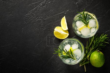 Téléchargez les photos : Gin tonique, cocktail traditionnel avec gin, tonique, glaçons, citron vert et romarin sur fond noir. Vue du dessus avec espace pour le texte. - en image libre de droit