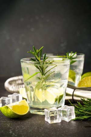 Foto de Gin tonic, cóctel tradicional con ginebra, tónico, cubitos de hielo, lima y romero sobre fondo negro. - Imagen libre de derechos