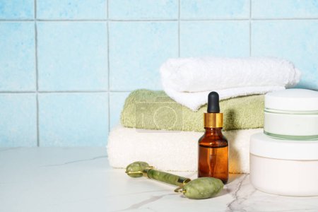 Foto de Productos para el cuidado de la piel en el baño. Crema facial, botella de suero, rodillo de jade y pila de toallas. - Imagen libre de derechos