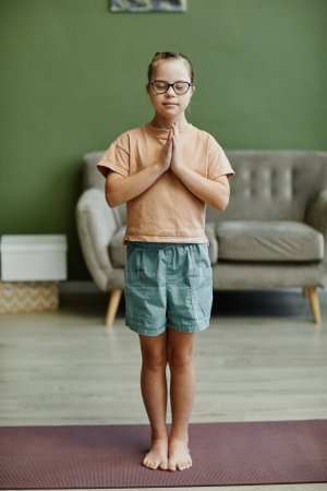 Téléchargez les photos : Vertical full length portrait of cute girl with down syndrome enjoying yoga at home while standing on mat eyes closed - en image libre de droit