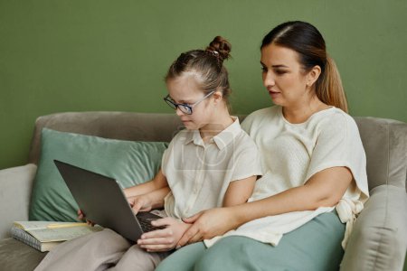 Photo for Portrait of mother and daughter with down syndrome relaxing on couch together in cozy home and using laptop, copy space - Royalty Free Image