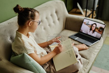 Photo for High angle portrait of young girl with down syndrome studying online at home while sitting on cozy couch and listening to tutor on screen - Royalty Free Image