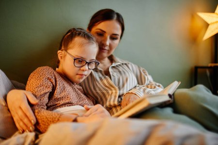 Photo for Portrait of little girl with down syndrome reading book at bedtime and relaxing with caring mother embracing her - Royalty Free Image