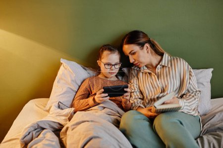Photo for Minimal portrait of caring mother and daughter with down syndrome using tablet to watch cartoons at bedtime, copy space - Royalty Free Image