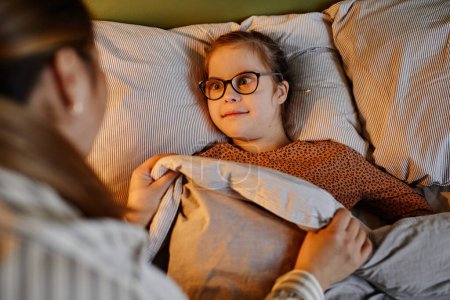 High angle portrait of caring mother tucking in child with down syndrome at bedtime in cozy lamp light, copy space
