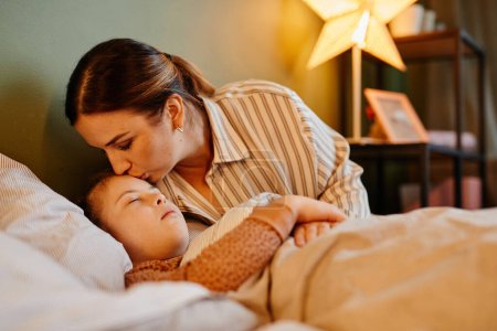 Photo for Portrait of caring mother kissing child with down syndrome at bedtime lit by cozy lamp light, copy space - Royalty Free Image