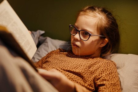 Photo for Portrait of young girl with down syndrome reading book in bed at night lit by cozy lamplight - Royalty Free Image