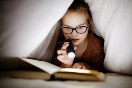 Téléchargez les photos : Front view portrait of young girl with down syndrome reading book under covers using flashlight, copy space - en image libre de droit