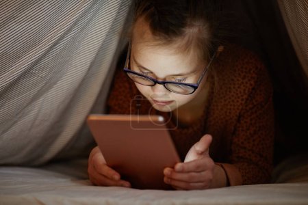 Téléchargez les photos : Portrait of young girl with down syndrome using smartphone in bed hiding under covers, digital obsession - en image libre de droit