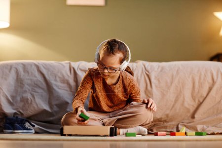 Foto de Portrait of young girl with down syndrome playing with toy blocks alone and wearing noise cancelling earphones, overstimulation concept, copy space - Imagen libre de derechos