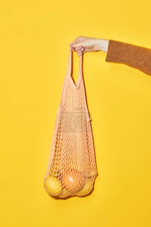 Foto de Vibrante primer plano de la mano sosteniendo eco bolsa de compras con naranjas sobre fondo amarillo, consumo consciente y concepto vegano - Imagen libre de derechos