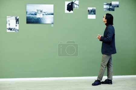 Photo for Young Asian man with long hair wearing eyeglasses spending time in modern art gallery looking at black and white photos on green wall at exhibition - Royalty Free Image