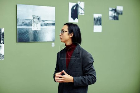 Téléchargez les photos : Critique d'art professionnel sérieux portant des lunettes debout dans la galerie d'art moderne regardant des photos en noir et blanc sur le mur à l'exposition - en image libre de droit
