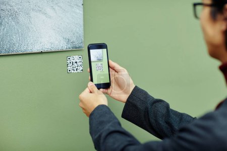 Téléchargez les photos : Jeune homme méconnaissable passer du temps dans la galerie d'art moderne numérisation QR code à l'aide de son smartphone pour obtenir plus d'informations sur la photo sur le mur - en image libre de droit