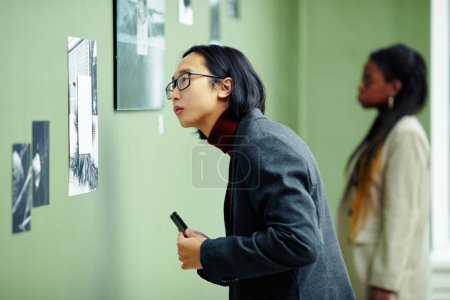 Téléchargez les photos : Portrait de beau jeune homme asiatique portant des lunettes visitant l'exposition dans la galerie d'art mettant l'accent sur la photo en noir et blanc sur le mur - en image libre de droit