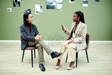 Téléchargez les photos : Femme afro-américaine assise sur une chaise tenant un enregistreur de voix interviewant un jeune photographe asiatique moderne le premier jour d'exposition dans une galerie d'art - en image libre de droit