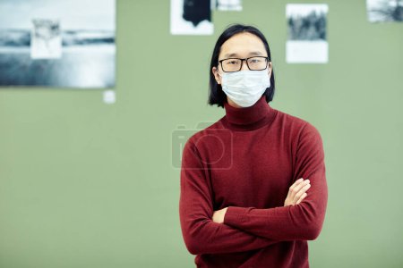 Foto de Joven hombre asiático con gafas y máscara protectora en la cara de pie con los brazos cruzados en la galería de arte moderno mirando a la cámara - Imagen libre de derechos