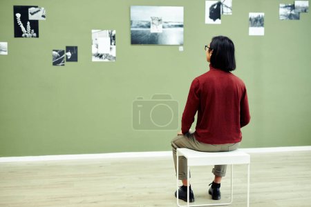 Téléchargez les photos : Vue arrière d'un jeune homme méconnaissable assis sur un tabouret dans une galerie d'art moderne regardant des photos en noir et blanc sur un mur vert - en image libre de droit