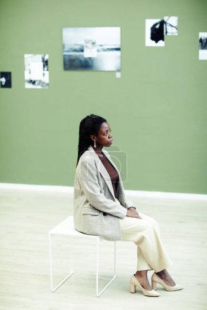Foto de Vista vertical de alto ángulo de la elegante joven mujer negra sentada en el taburete en la galería de arte contemporáneo mirando fotos en la pared en la exposición - Imagen libre de derechos