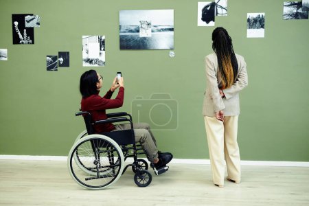 Photo for Unrecognizable young Asian man with disability and stylish Black woman visiting contemporary photography exhibition in art gallery looking at photos and scanning qr code - Royalty Free Image
