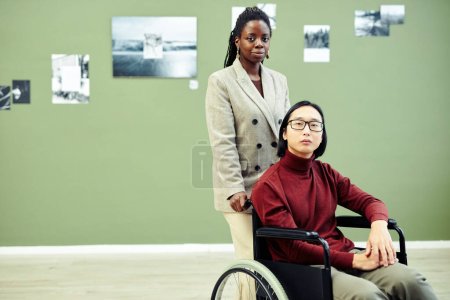 Téléchargez les photos : Jeune femme noire élégante et son amie asiatique handicapée en fauteuil roulant visitant l'exposition de photographie contemporaine dans la galerie d'art - en image libre de droit