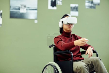 Photo for Horizontal medium shot of young Asian man with disability wearing VR headset visiting exhibition in modern art gallery using augmented reality technology - Royalty Free Image