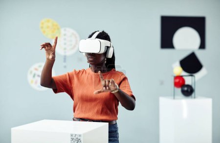 Foto de Mujer afroamericana joven con auriculares VR mirando objetos de arte en exposición contemporánea usando tecnología de realidad aumentada - Imagen libre de derechos