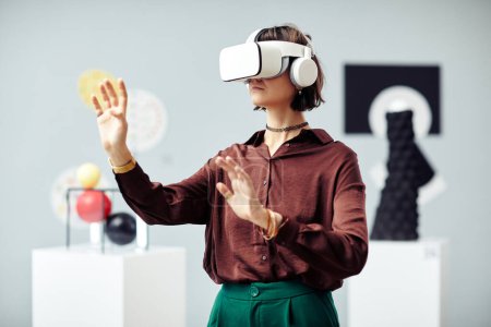 Foto de Mujer caucásica joven con auriculares VR mirando objetos de arte mientras visita la exposición moderna utilizando tecnología de realidad aumentada - Imagen libre de derechos