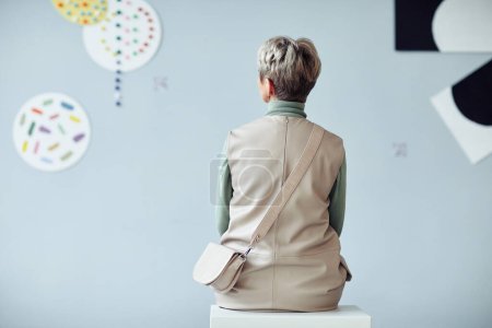 Téléchargez les photos : Vue arrière d'une femme mûre méconnaissable assise sur un tabouret dans une galerie d'art moderne regardant des œuvres d'art - en image libre de droit