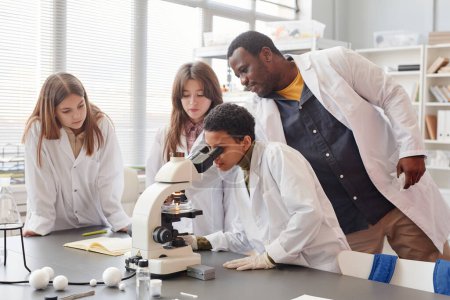 Téléchargez les photos : Vue de côté portrait de jeune fille noire regardant au microscope avec tout en faisant des expériences avec un groupe d'enfants dans le laboratoire de chimie de l'école, espace de copie - en image libre de droit