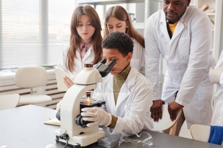 Téléchargez les photos : Portrait de jeune fille noire regardant au microscope tout en faisant des expériences avec un groupe d'enfants dans le laboratoire de chimie de l'école - en image libre de droit
