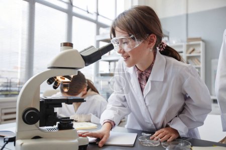 Téléchargez les photos : Portrait de jeune fille souriante regardant au microscope tout en appréciant les expériences dans le laboratoire de chimie de l'école - en image libre de droit
