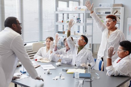 Téléchargez les photos : Groupe diversifié d'enfants excités levant la main tout en répondant à des questions en classe de chimie à l'école - en image libre de droit