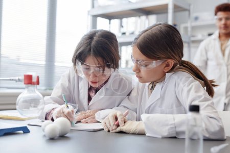 Téléchargez les photos : Portrait de deux filles prenant des notes dans un carnet tout en faisant des expériences scientifiques dans un laboratoire de chimie à l'école - en image libre de droit