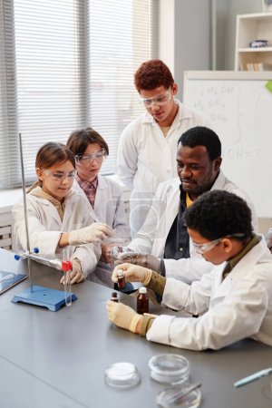 Foto de Retrato vertical del profesor afroamericano demostrando experimentos científicos a un grupo de niños en el laboratorio de química de la escuela - Imagen libre de derechos