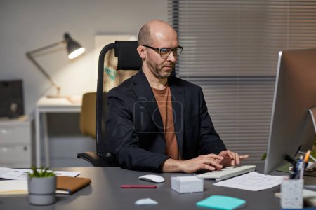 Foto de Retrato del hombre de negocios adulto calvo que usa la computadora en el lugar de trabajo mientras trabaja hasta tarde en la oficina - Imagen libre de derechos