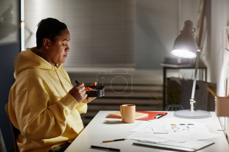 Photo for Side view portrait of Latin American woman eating takeout at desk while working late at night in office, copy space - Royalty Free Image