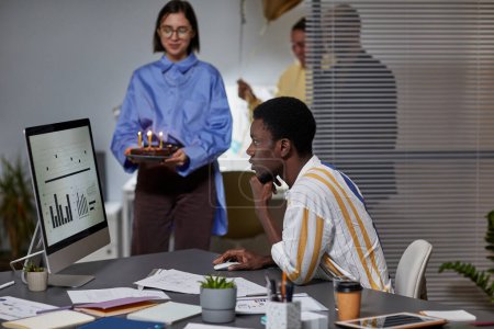 Téléchargez les photos : Vue de côté portrait de l'homme afro-américain travaillant tard dans le bureau avec des collègues apportant gâteau d'anniversaire en arrière-plan - en image libre de droit