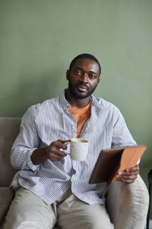 Téléchargez les photos : Portrait vertical minimal d'un bel homme afro-américain tenant une tasse de café et regardant la caméra tout en se relaxant sur un canapé contre un mur vert - en image libre de droit