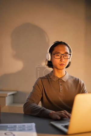 Téléchargez les photos : Portrait minimal de jeune homme asiatique portant un casque sur le lieu de travail tout en travaillant tard dans le bureau éclairé par des lampes - en image libre de droit